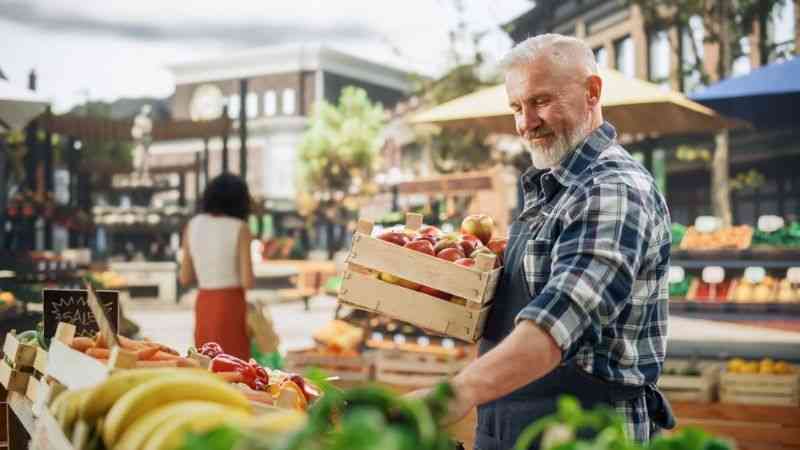 A middle-aged white male with short grey hair and a trimmed white beard, wears a blue and white plaid shirt under a navy apron. He is stood in front of a fresh fruit and vegetable market stall, carrying a wooden crate of apples, smiling proudly as he tends to his display of quality goods, supporting the community with NatWest.
