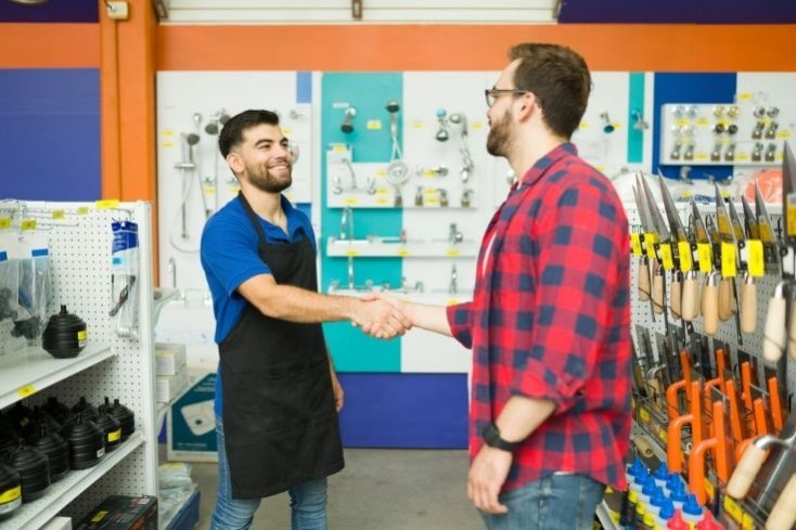 Interior of a hardware business, with tools and plumbing products on display. The young business owner, white male with short dark hair and a beard, wearing a blue polo shirt, black apron and jeans, smiles as he shakes hands with a young white male customer, who wears a red and blue check shirt and jeans, has short brown hair, a beard and glasses.