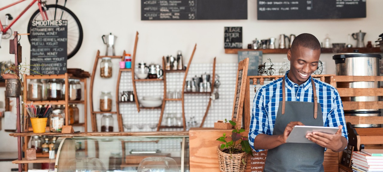A young black café owner smiles, operating a tablet, representing the success brought by NatWest's business  support for ethnic minorities.