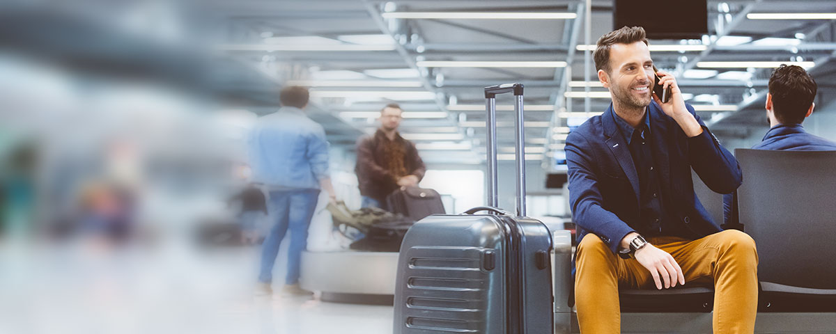 A young white male business plus traveller with short dark hair, dressed in a navy jacket, navy shirt, and beige trousers, is sat in a modern, light, airport departure lounge, smiling, relaxed as he talks on his phone, a large silver luggage case beside him.