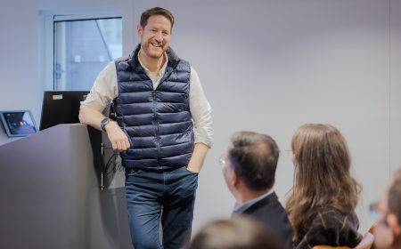 David Grunwald wearing a gilet, standing and speaking to a room full of people. 