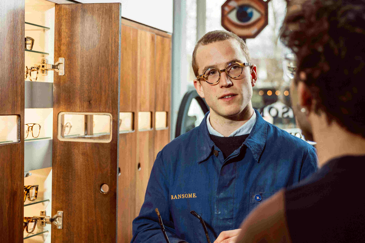 Jack, a young white male, business owner of Ransome Optical, has cropped ginger hair, wears bespoke tortoise shell framed glasses and navy workwear embroidered with his brand. He stands in his eyewear shop, in conversation with a customer.