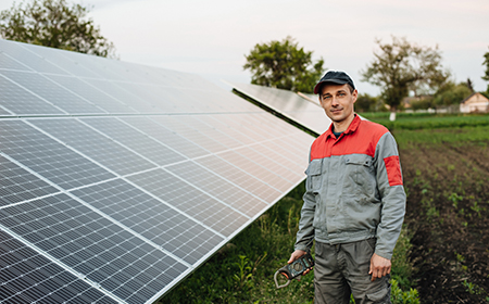 Young white male, wearing baseball cap and red/ grey jacket, stands in a field beside large solar panel installation holding digital measuring device.