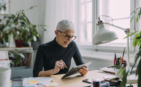 White female, white hair and black rimmed glasses, sits at a home desk by a window, wearing black long sleeve top, engrossed with tablet and pen.