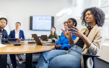 A woman talking whilst sat at a table in a meeting room.