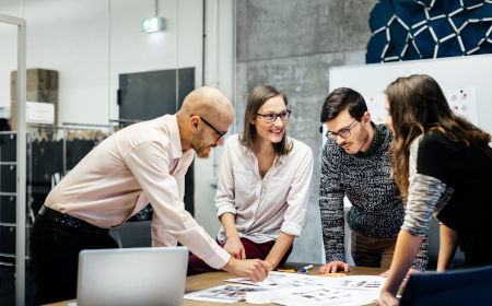 A group of people in a meeting room looking at some blueprints spread out on a table.