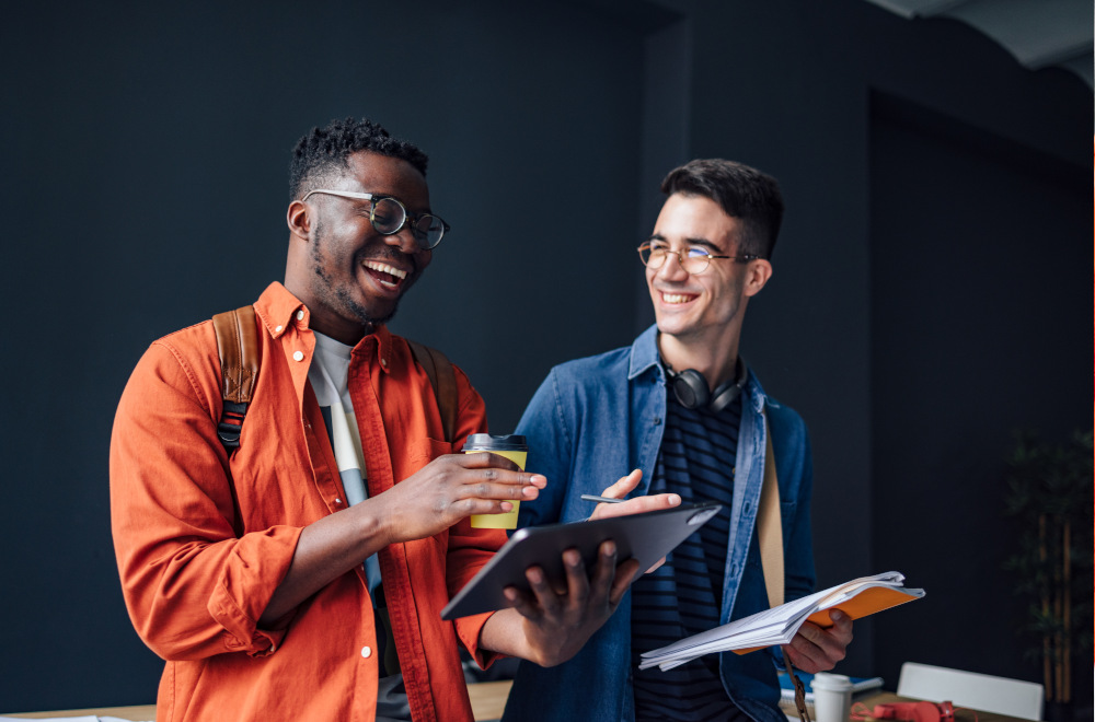 Two smiling men dressed casually and looking at tablets and a notebook whilst holding takeaway coffee cups.