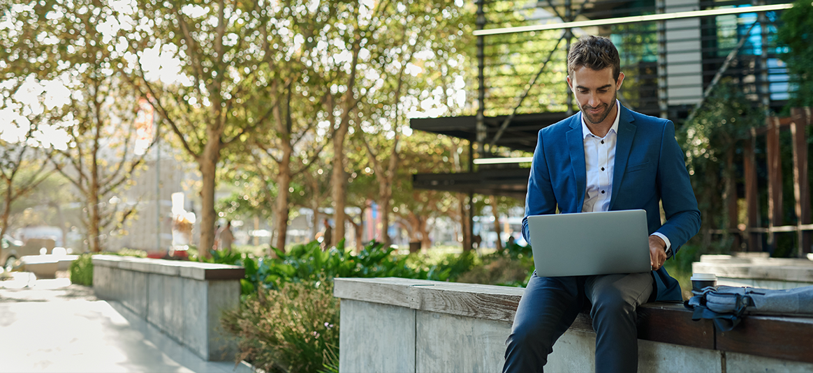 Photo of a suited man sitting on a park bench and looking at an open laptop.