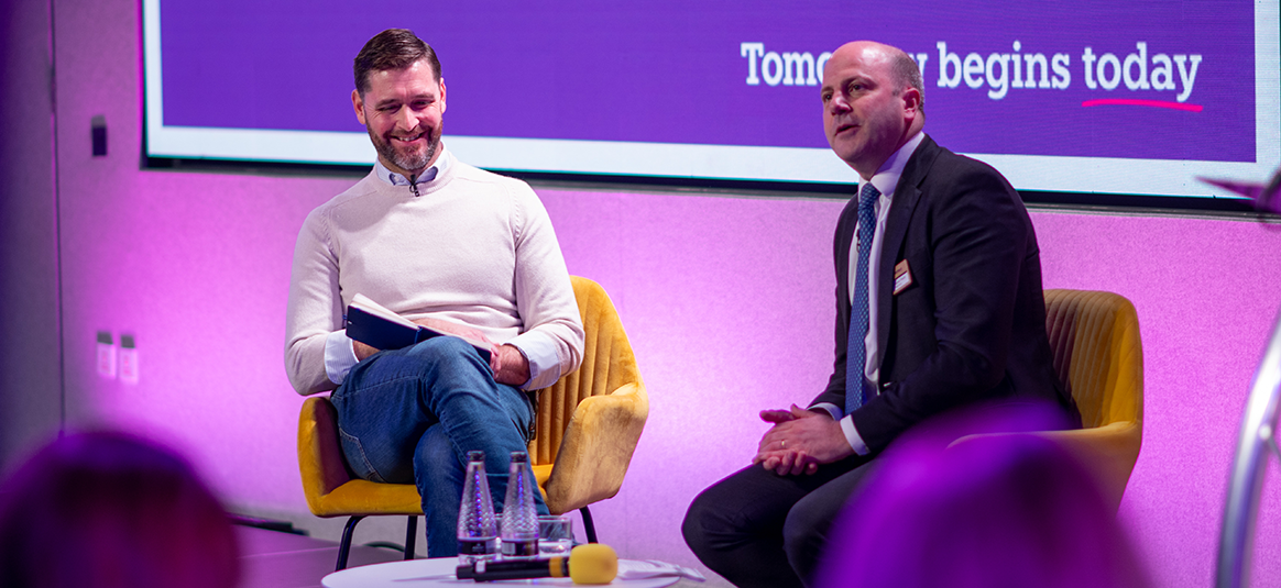 A professional event setting featuring two individuals seated on modern yellow chairs on a stage. One person is holding a notebook, while the other has hands resting on their lap. A small round table in front of them holds two water bottles, a microphone,