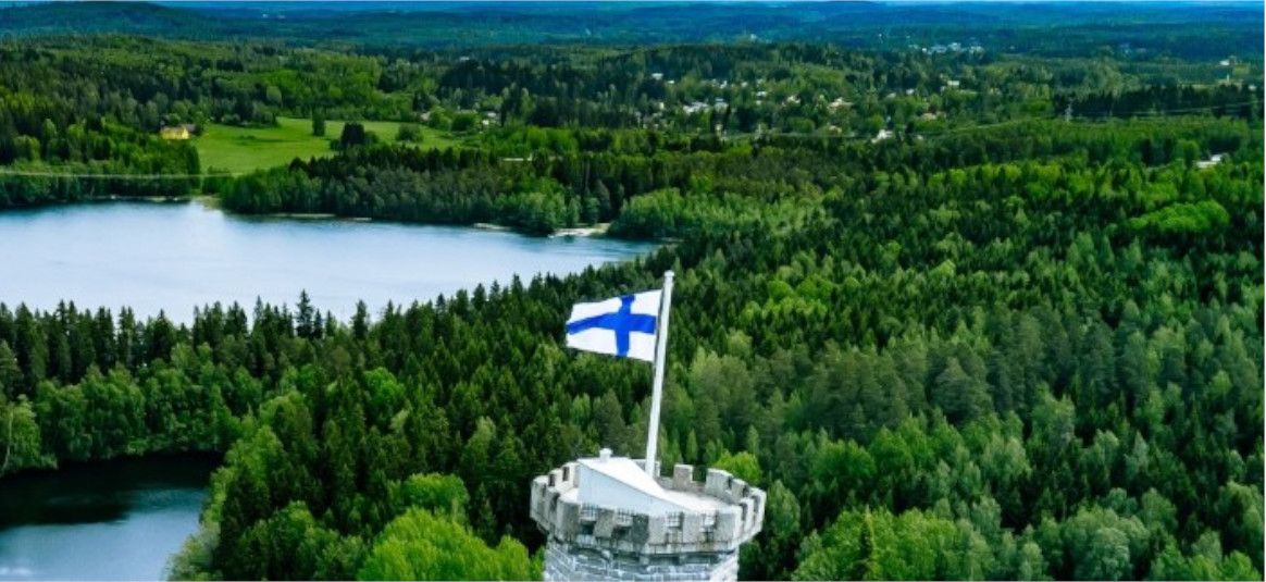 A castle flying the Finnish flag overlooks a lake in the middle of a forest.