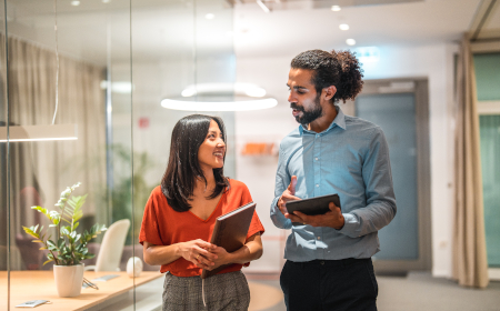 Two people walking in an office hallway talking and holding tablets.
