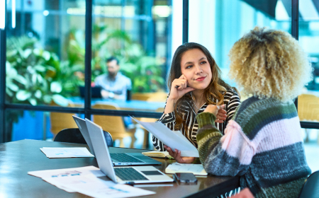 Two women sitting at a table in an office building with printouts and laptops.