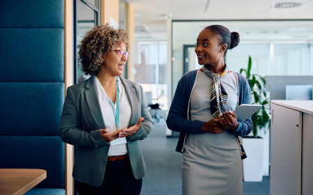 Two women in professional clothing smiling and talking as they walk across an open plan office