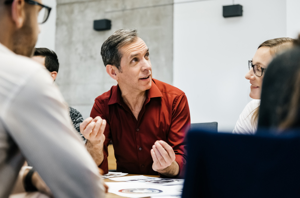A man wearing a red shirt and talking passionately to other people in a meeting room. 