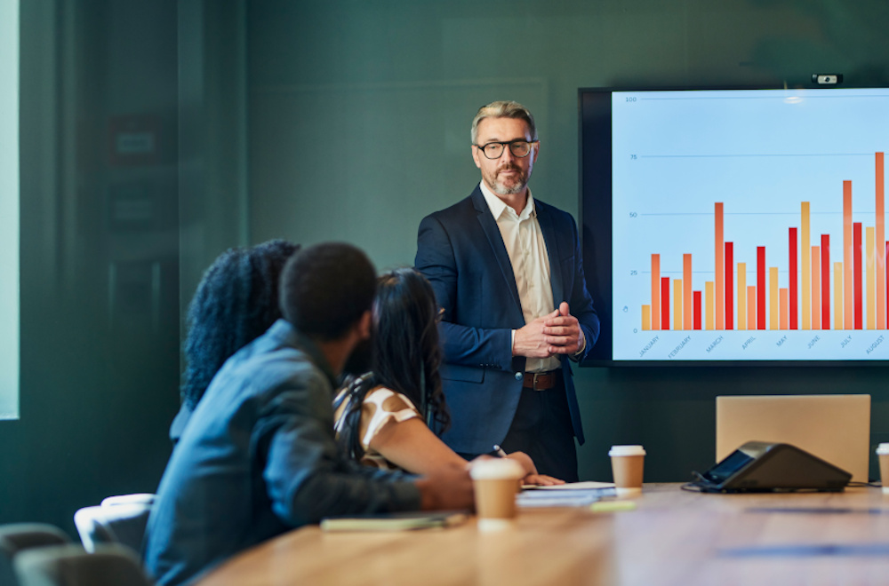 A man wearing glasses presenting to clients during a team meeting in an office setting.