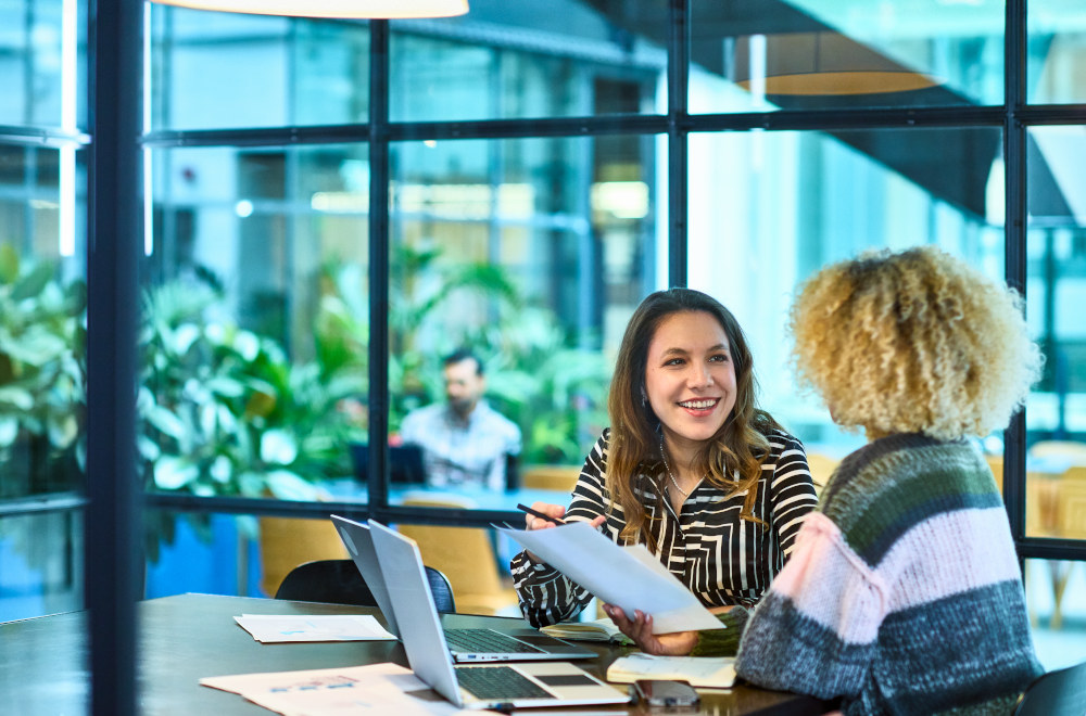 Two women working together on a laptop and discussing a project in a modern office setting.