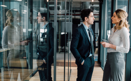 Two people standing and talking in a glass-lined office hallway.