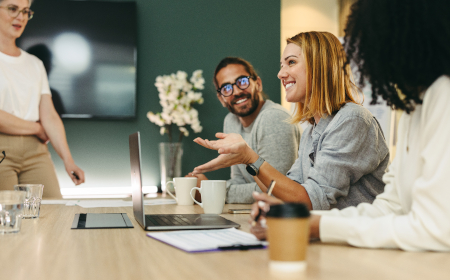 Three smiling people sitting at an office desk in front of an open laptop.