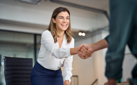 A professionally dressed woman in an office environment shaking a colleague's hand.