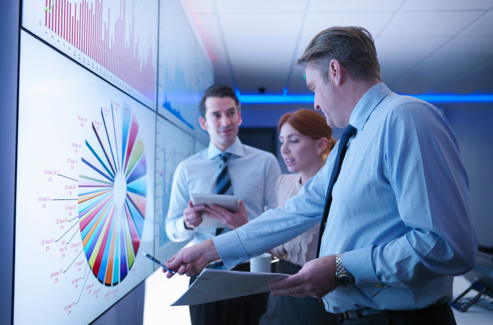 Three people in a professional office environment viewing a wall projection displaying data charts and numerical figures.