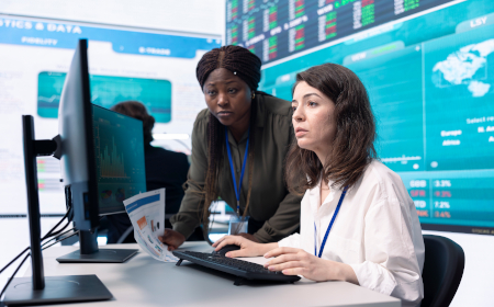 Two women in an office environment looking at data on a computer with stocks on a board behind them.