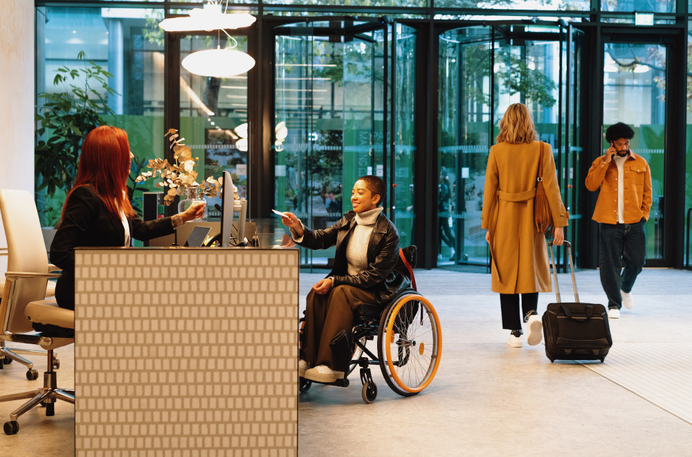 A woman in a wheelchair waiting at a hotel reception with people wheeling luggage as they walk to the door.