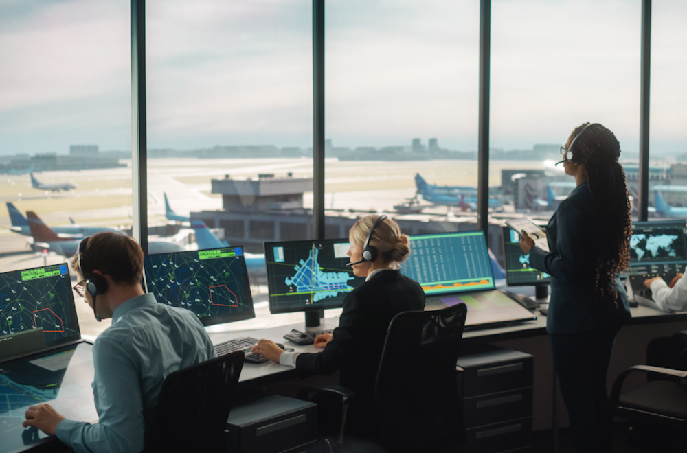 Employees in an airport control tower looking out the window at the runway below them.