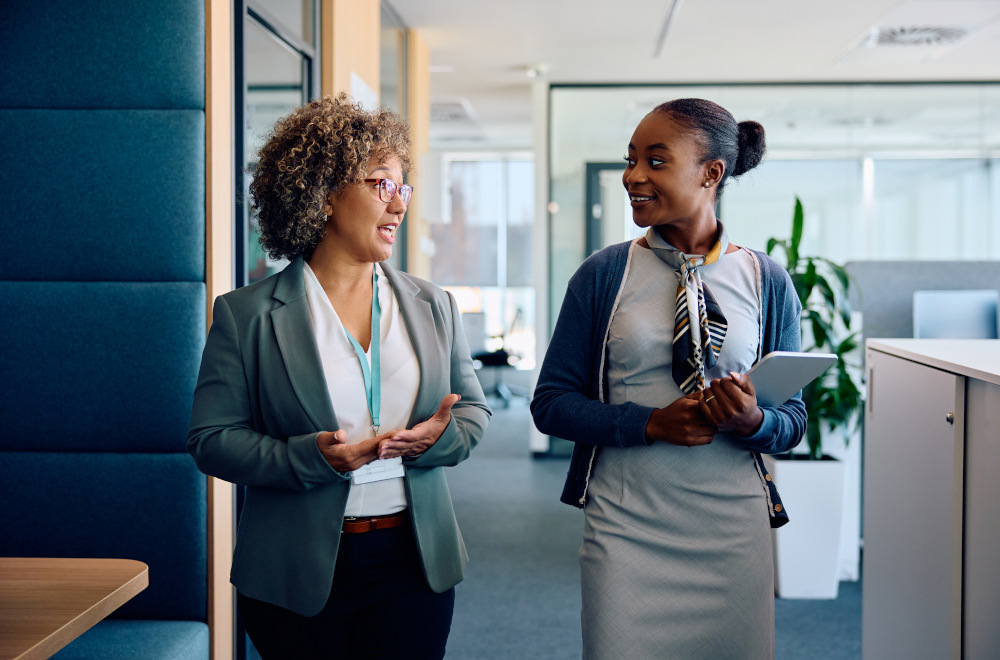 Two professionally dressed women walking and talking in an office setting, with one holding an iPad.