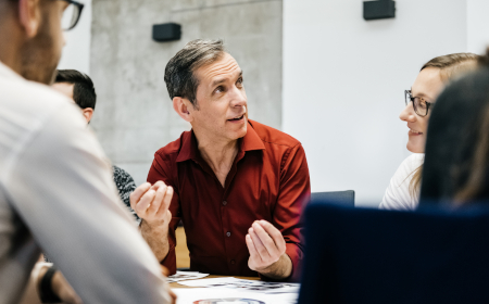 A man wearing a red shirt sat at a meeting table talking to his colleagues.