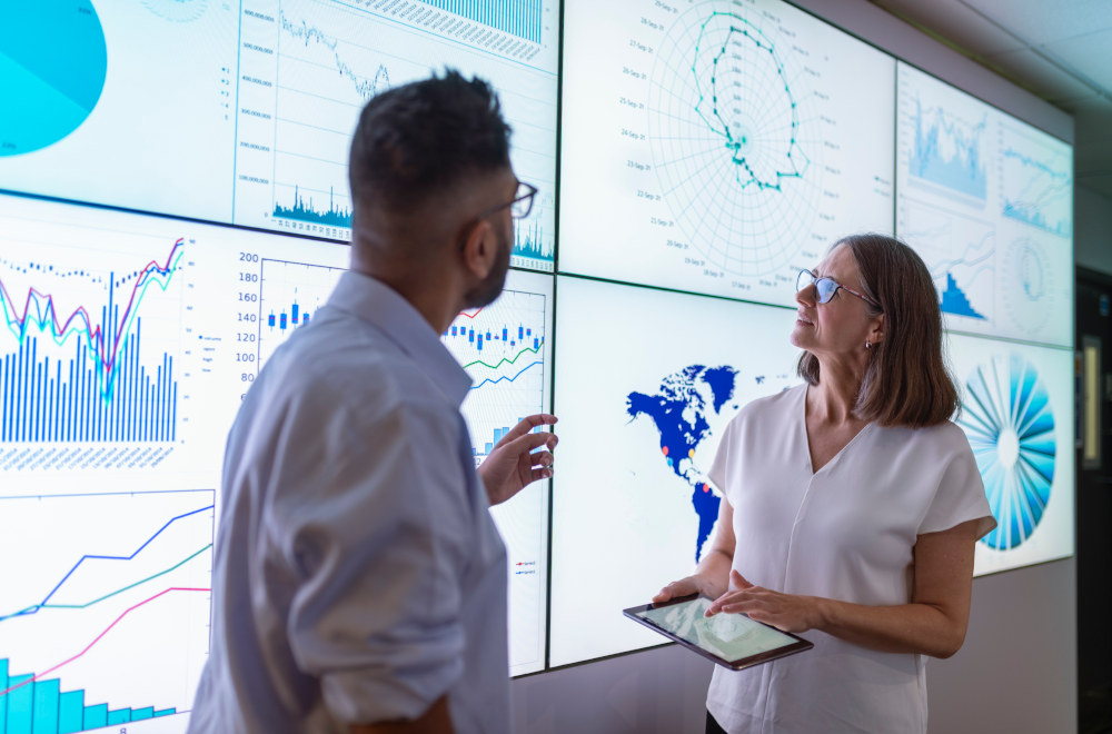 Two professional colleagues in an office setting analyzing data charts displayed across multiple screens.
