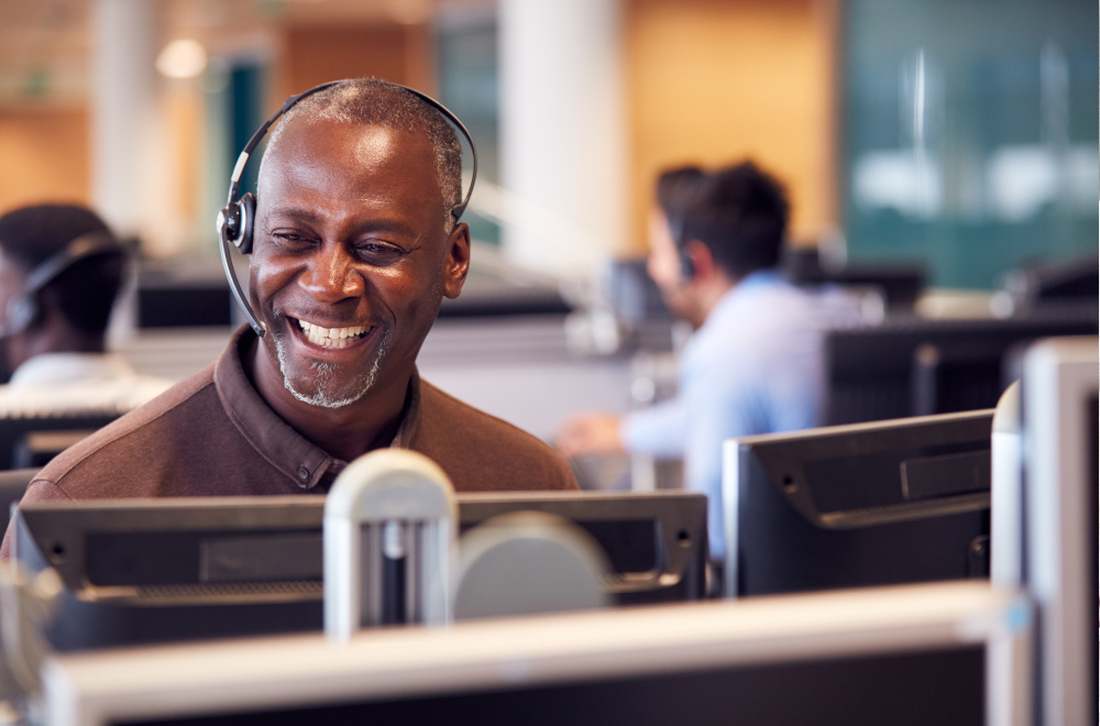A man in a call centre wearing a headset and smiling at a computer.