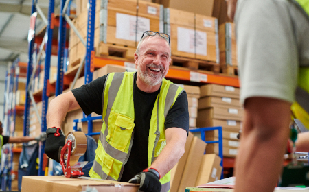 A smiling man in a warehouse wearing a hi vis jacket holding a cardboard box.