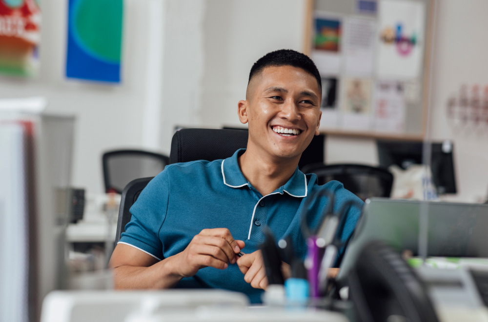 A man in an office environment smiling at a computer.