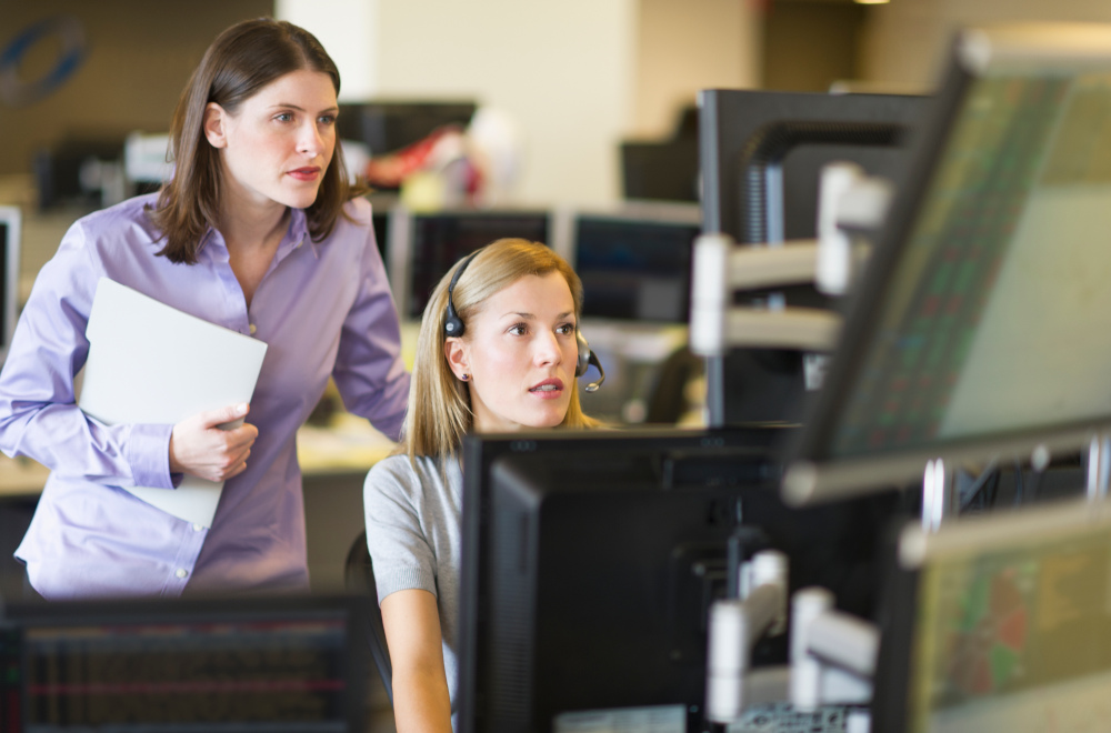 Two women sat in front of computers, one wearing a headset, and looking at the screen.