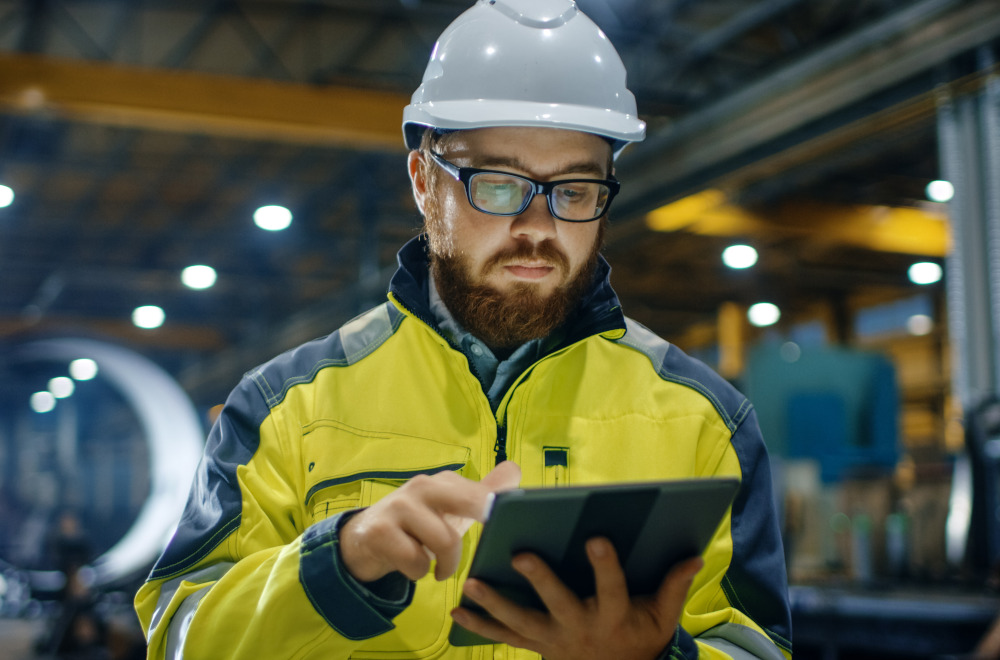 A man wearing a hard hat, glasses and a hi-vis jacket looking at the tablet that he is holding.