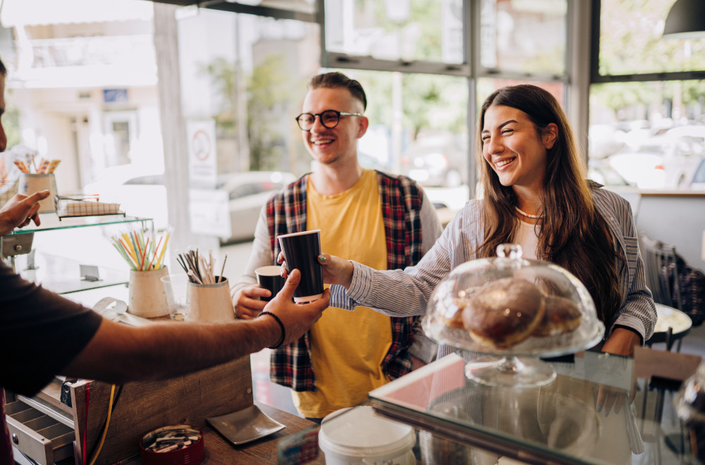 A man and a woman in a coffee shop being handed a takeaway cup of coffee by a barista.