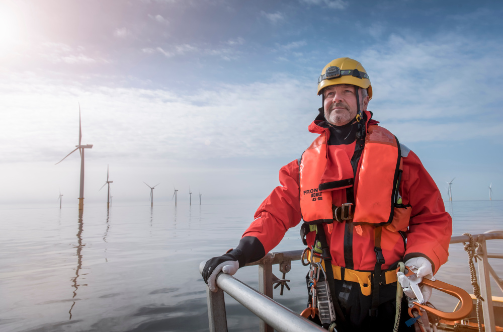 A sea engineer wearing safety gear stands on the deck of a boat, with wind turbines visible in the background under a blue sky.