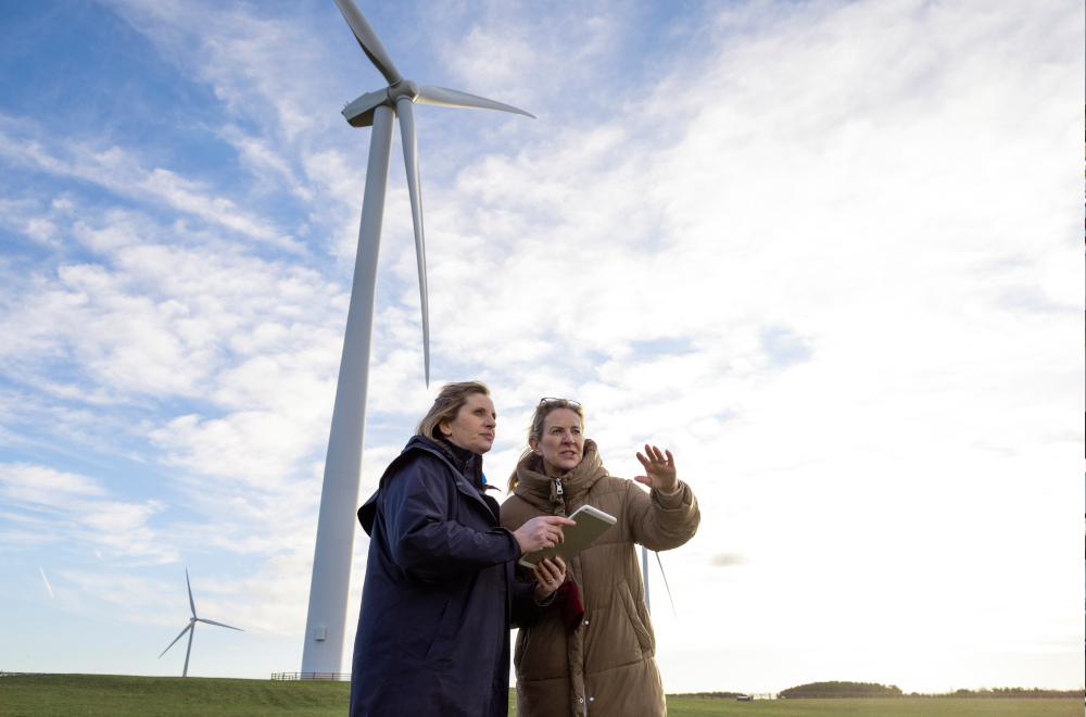 Two people dressed in warm coats standing in a field with wind turbines.