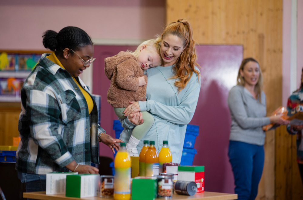 A woman holding her young child chatting to another woman who is putting squash and canned goods onto a table.