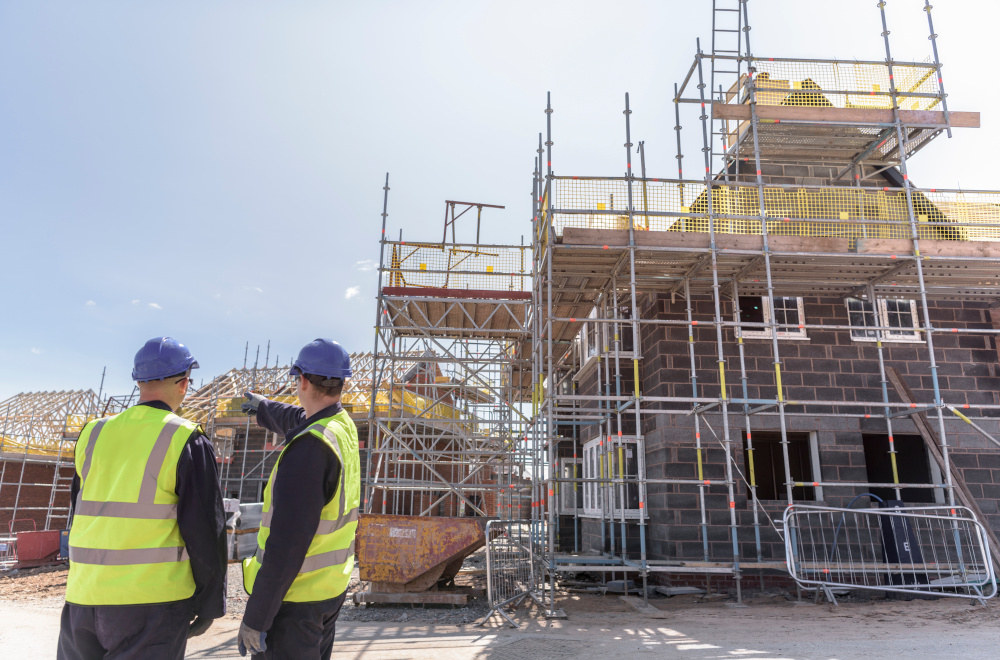 Two workers wearing hi vis and hard hats looking at a part built house with scaffolding.