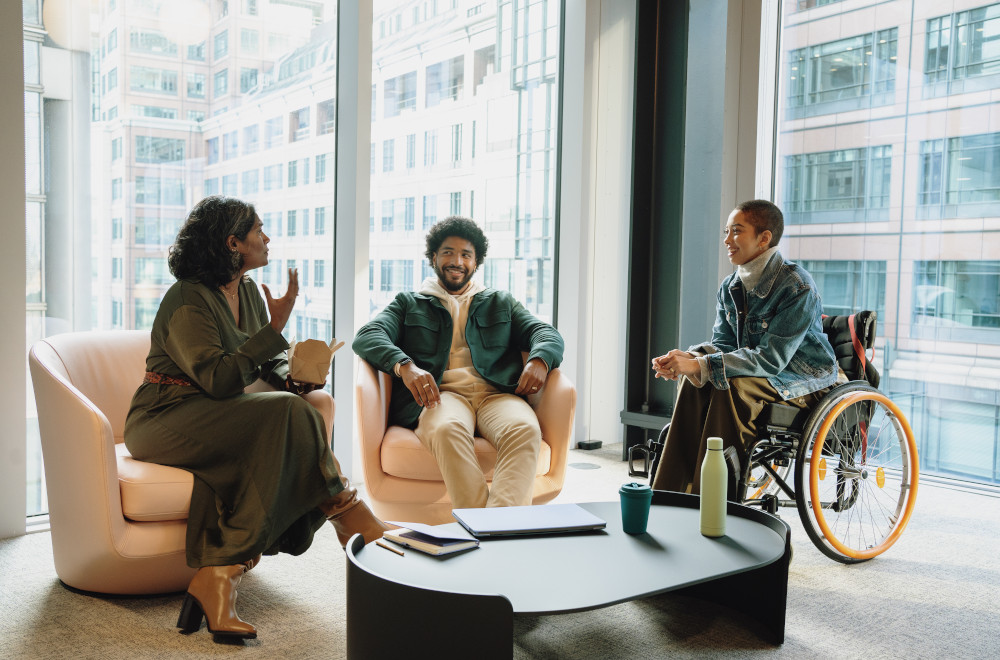 Three business professionals, including one in a wheelchair, sitting together in an office setting and collaborating, reflecting inclusivity and teamwork in a modern workplace.